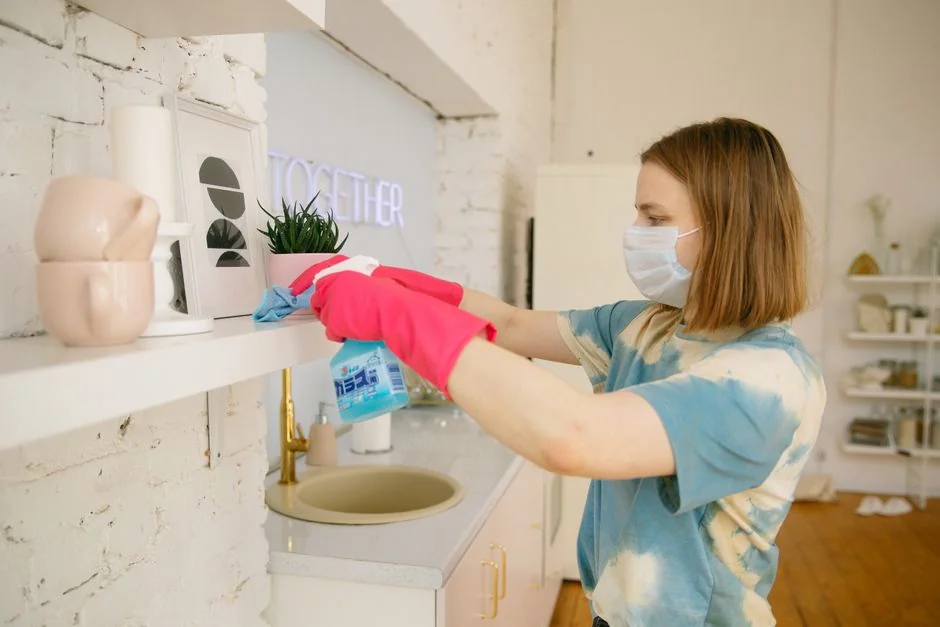 A hand wearing a yellow rubber glove holds a red and white spray bottle against a neutral background.